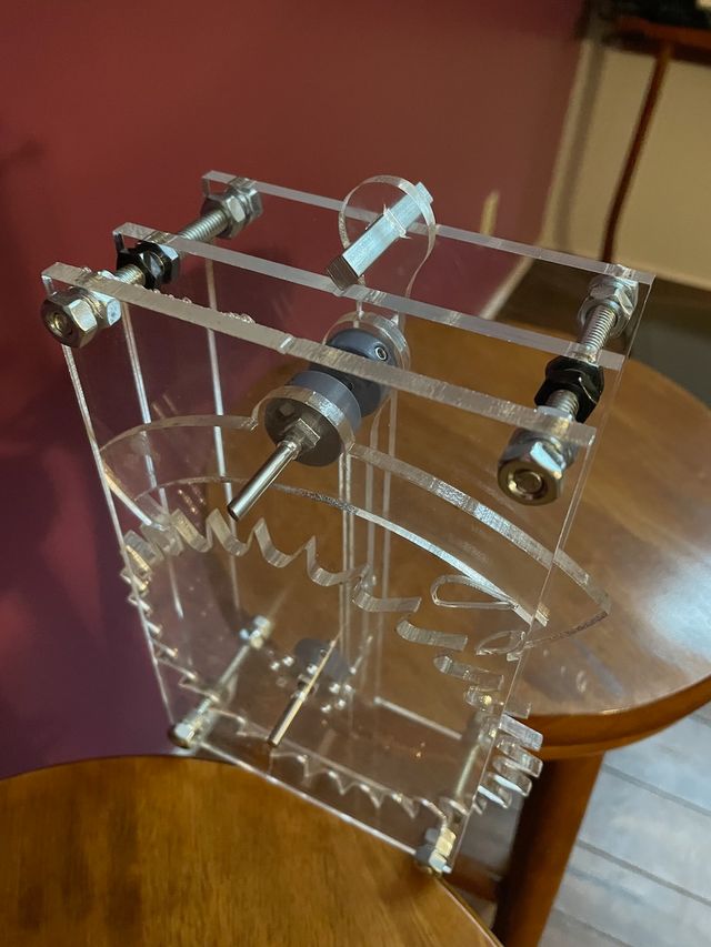 Prototype of a clear acrylic mechanical clock movement, just the pendulum and escape mechanisms, sitting across two stools so the pendulum can hang between
