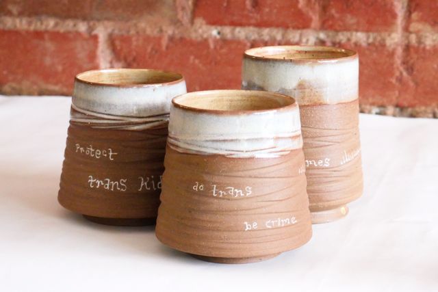 Three tumblers, also wide base with grooved walls sloped in - but the clay is brown un-speckled, the lips are an icy white, and they have hand-written text. The front one says "do trans, be crime". We can also see the end of "…crimes" sticking out on one behind, and "protect trans kids" on the last one