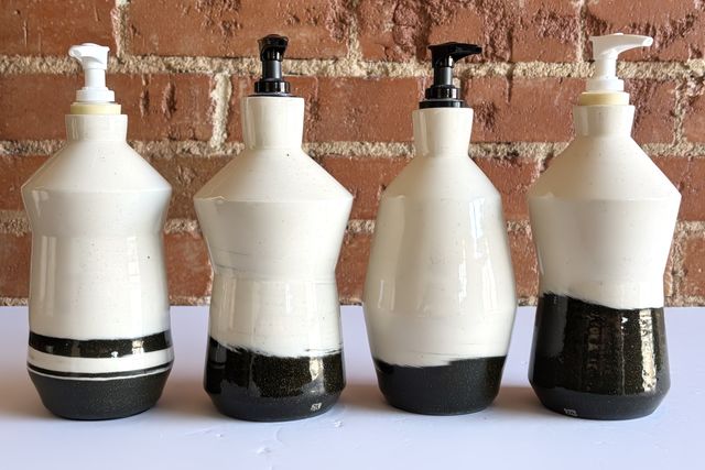 A set of angular ceramic bottles, some with an inset hourglass waist, some outset and diamond-shaped. All of them have a black clay base and a hard slightly-swirled transition to white clay for the upper two-thirds. They also have soap dispenser pumps in the top.
